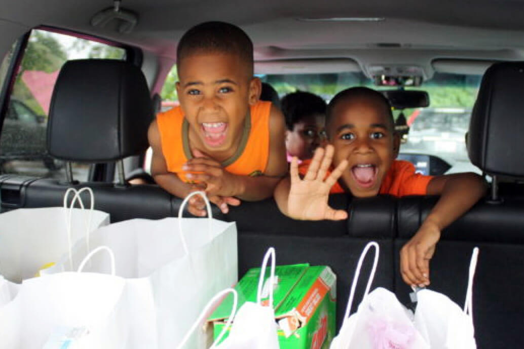 Boys in the backseat of a car with food in the back hatch, looking out the back, smiling at a camera.